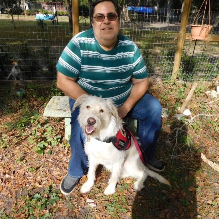 Man in late 50s with service dog sitting on a stone bench outdoors, smiling at the camera in a garden setting.